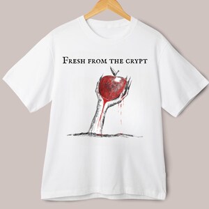 Può includere: T-shirt bianco con la scritta "Fresh From The Crypt". La maglietta presenta una grafica di una mano che emerge dal terreno, che tiene una mela rossa. La mela e la mano hanno un aspetto sanguinolento, con gocce rosse.