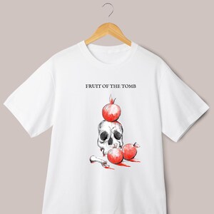 Può includere: Maglietta bianca con un'illustrazione in bianco e nero di uno scheletro con una melagrana rossa in cima. Il testo "FRUIT OF THE TOMB" è stampato sopra lo scheletro.