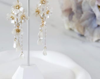 May include: A pair of white flower dangle earrings with gold accents and pearl beads hanging from a chain. The earrings are displayed on a white stand.