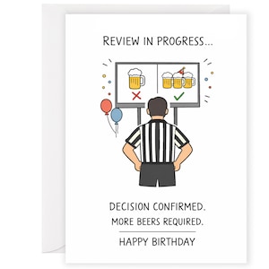 May include: A white greeting card featuring a cartoon referee in a black and white striped shirt looking at a screen showing beer mugs. Text reads: REVIEW IN PROGRESS... DECISION CONFIRMED. MORE BEERS REQUIRED. HAPPY BIRTHDAY. Red and blue balloons.