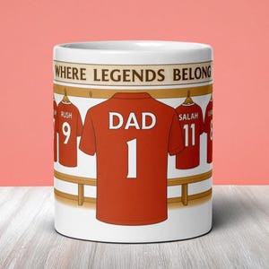 May include: White ceramic mug featuring a red jersey with "DAD 1" on the back, surrounded by other jerseys. The mug has the text "WHERE LEGENDS BELONG" at the top. A great gift for any dad.