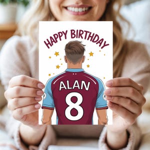 May include: A white card with the words "Happy Birthday" in a circle around a cartoon image of a soccer player wearing a maroon and blue jersey with the name "ALAN" and the number "8" on the back. The background is white with gold stars.