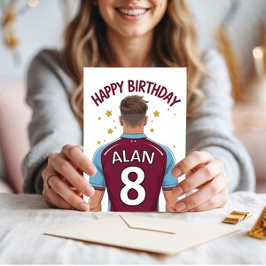 May include: Happy Birthday card featuring a cartoon illustration of a person wearing a maroon and blue soccer jersey with the name "ALAN" and the number "8" on the back. The card has a white background with gold stars.