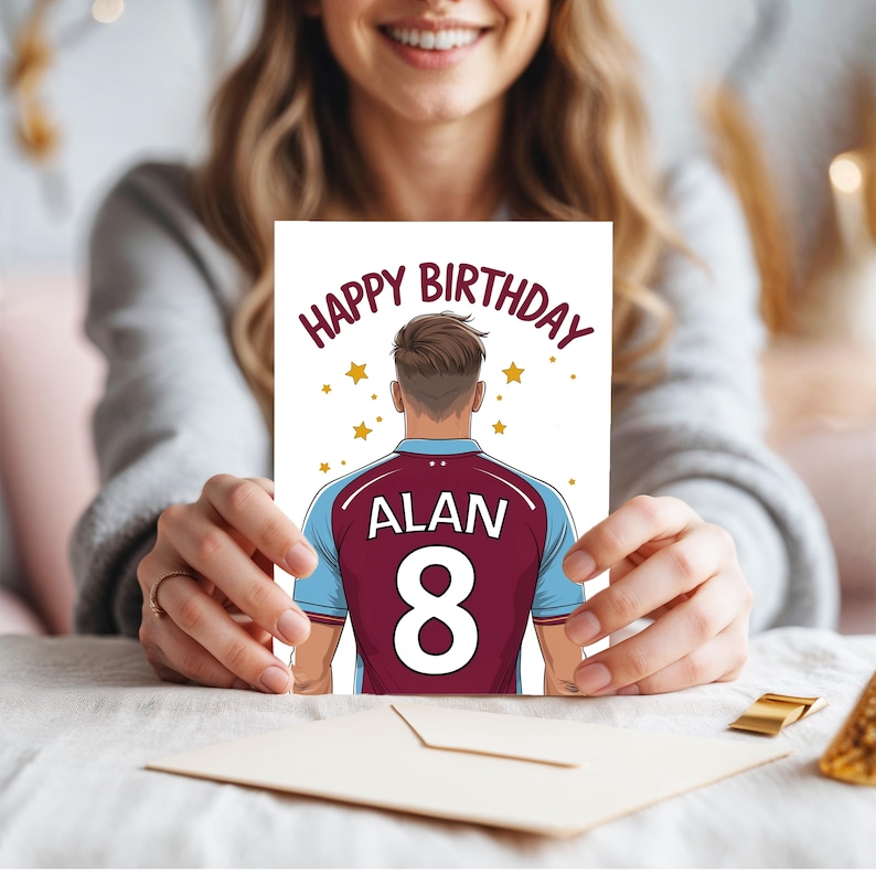 May include: Happy Birthday card featuring a cartoon illustration of a person wearing a maroon and blue soccer jersey with the name "ALAN" and the number "8" on the back. The card has a white background with gold stars.