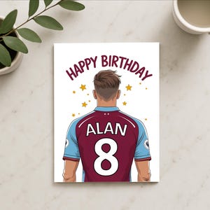 May include: A white card with the text "Happy Birthday" in a gold font. The card features a cartoon illustration of a person wearing a maroon and blue soccer jersey with the name "Alan" and the number "8" on the back. The background is white with gold stars.