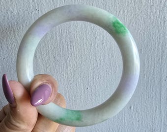 May include: A jade bangle bracelet with a smooth, polished surface. The bracelet features a blend of colors, including white, green, and lavender. The jade is held against a light gray background.