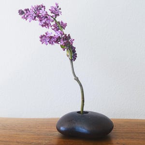May include: A minimalist floral arrangement featuring a dark, pebble-shaped vase holding a sprig of purple lilacs. The vase sits on a wooden surface against a white backdrop, creating a serene and natural aesthetic.