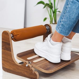 Könnte beinhalten: Ein Fußhocker aus Holz mit einer braunen gepolsterten Fußstütze und einer braunen Holzplattform. Eine Person trägt weiße Turnschuhe und Jeans und benutzt den Fußhocker.