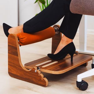 Op de afbeelding: Een houten voetensteun met een gebogen ontwerp en een oranje, gewatteerde rol. De voetensteun heeft een platform voor de voeten en is ontworpen om ergonomische ondersteuning te bieden. Het product is gemaakt van hout met een natuurlijke afwerking.