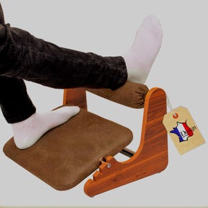 Peut inclure: Un repose-pieds en bois avec une plateforme rembourrée marron et un support en bois incurvé. Le repose-pieds porte une étiquette indiquant "Design en France". La personne sur l'image porte un pantalon noir et des chaussettes blanches.