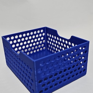 Puede incluir: Una cesta de almacenamiento rectangular azul con un diseño perforado. La cesta tiene esquinas redondeadas y una abertura para el asa en un lado. La cesta está hecha de un material sólido y se encuentra sobre una superficie blanca.