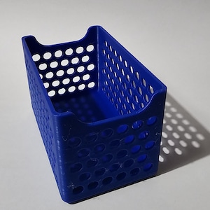 Può includere: Un piccolo contenitore blu rettangolare con angoli arrotondati e un design perforato. Il contenitore ha una maniglia su ciascuna estremità ed è realizzato in un materiale solido e opaco. Il contenitore è vuoto e poggia su una superficie bianca.