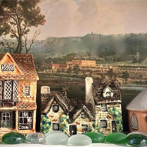 May include: A collection of ceramic miniature houses with detailed facades, including a bookstore and a coin shop, set against a landscape painting. Green and clear glass pebbles are in the foreground.
