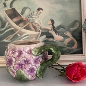 May include: A ceramic mug with a floral design featuring pink and purple flowers and green leaves. The mug has a handle shaped like a leaf.