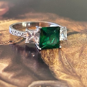 May include: A silver ring featuring a large, square-cut emerald green gemstone flanked by two smaller, square, clear gemstones. The band is detailed with small, clear stones. The ring is set against a blurred, artistic background.
