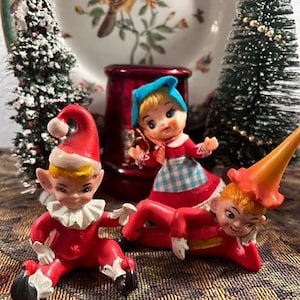 May include: A festive display featuring three vintage Christmas figurines. Two elves in red suits and hats are positioned near a doll in a blue bonnet and red dress. The background includes miniature Christmas trees and a decorative plate.
