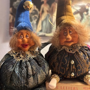 May include: Two whimsical fabric dolls with expressive faces. One doll wears a blue hat with a gold bell, the other a tan hat. Both dolls have textured hair, ornate jackets, and gold trim. They sit atop a book with the title partially visible.
