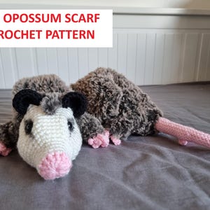 May include: A handmade crochet opossum scarf in shades of grey, white, black, and pink. The scarf is designed to resemble a 3D opossum. The text "3D Opossum Scarf Crochet Pattern" is displayed in red and white.