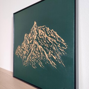 May include: A framed canvas art piece featuring a gold mountain range design against a dark green background. The artwork is signed in the lower right corner and is encased in a black frame. The mountain range is textured.