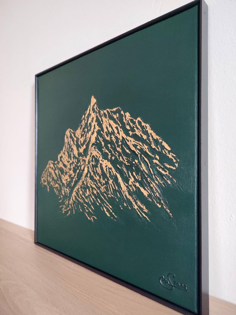 May include: A framed canvas art piece featuring a gold mountain range design against a dark green background. The artwork is signed in the lower right corner and is encased in a black frame. The mountain range is textured.