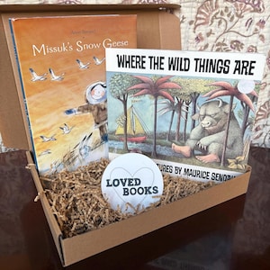 May include: A cardboard box filled with brown packing paper and two children's books. The books are "Missuk's Snow Geese" by Anne Renaud and "Where the Wild Things Are" by Maurice Sendak. A white sticker with the text "LOVED BOOKS" is on top of the packing paper.