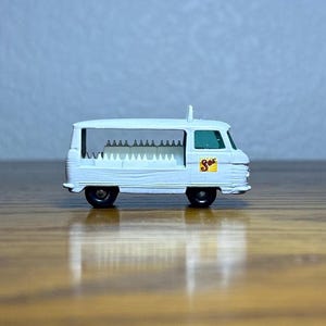 Puede incluir: Camión de reparto de juguete blanco con un parabrisas verde y un logotipo "So" amarillo y rojo en el lateral. El camión lleva una carga completa de botellas de leche de juguete blancas.