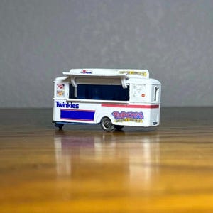 May include: A white toy food truck with a blue and white "Twinkies" logo on the side and a pink and white "Pop-Tarts" logo on the side. The truck has a white roof and a blue and white striped awning.
