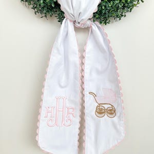 Könnte beinhalten: Weißes Babytuch mit rosa gewellten Kanten und gestickten Details. Mit einem rosa Monogramm und einem Kinderwagendesign in Gold und Rosa. Oben verknotet.