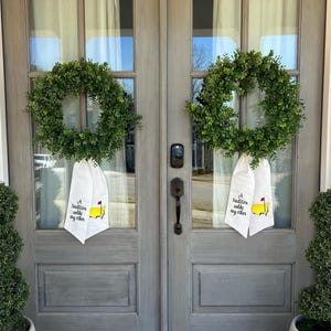 May include: Two wreaths made of green boxwood hang on a pair of gray double doors. Each wreath has a white towel hanging from it with the text "A tradition we share with each other" and a yellow and red golf course graphic.