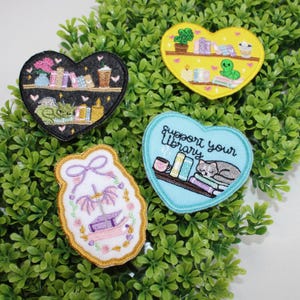 May include: Four embroidered patches with book-themed designs. One black heart patch features a dragon, books, and a coffee cup. A yellow heart patch shows books, a plant, and an alien. A blue heart patch says "Support your Library" with a cat. A white patch has a book.