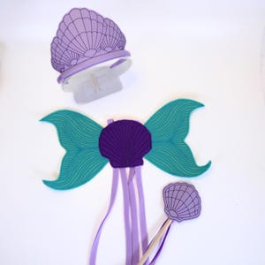 May include: A mermaid costume set in purple and green. The set includes a headband with a purple seashell crown, a pair of wings with a purple seashell in the centre, and a wand with a purple seashell on the end.