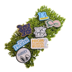 Może przedstawiać: Zbiór haftowanych naszywek z tekstami i wzorami z popularnych musicali i filmów. Naszywki obejmują "Beauty and the Beast", "Frozen", "High School Musical 2", "Broadway Bound", "13 The Musical", "Harvey" i "Wicked". Naszywki są ułożone na zielonym listowiu.