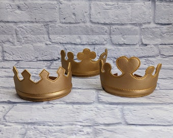May include: Three gold-colored crowns with different decorative tops are arranged on a light gray brick background. The crowns are made of a soft material and have a stitched design. The crowns are likely for a child's costume or party.