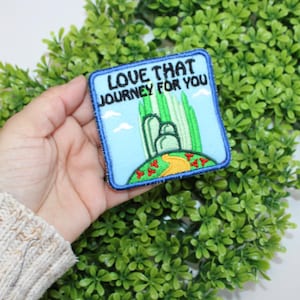 Könnte beinhalten: Quadratischer Aufnäher mit dem Text "LOVE THAT JOURNEY FOR YOU" über einer grün-gelben Illustration eines Weges, der zu einer grünen Bergkette führt. Der Aufnäher hat einen blauen Rand und ist vor einem Hintergrund aus grünem Laub angeordnet.