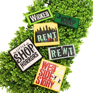 May include: A collection of six rectangular fabric patches featuring Broadway musical themes. The patches display the titles Wicked, Jungle Book, Rent, Little Shop of Horrors, and West Side Story in various colors, fonts, and graphic designs.