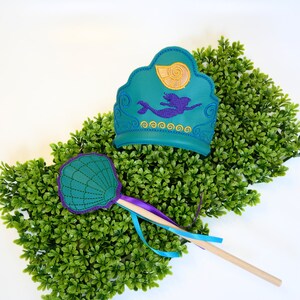 May include: Teal and purple mermaid-themed crown and wand set. The crown features a mermaid design and seashell, with gold and purple embroidery. The wand has a seashell design with purple trim and ribbons. The set is placed on a green, textured background.