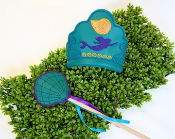 Puede incluir: Juego de corona y varita mágica con temática de sirena, en color turquesa y morado. La corona presenta un diseño de sirena y una concha, con bordados dorados y morados. La varita tiene un diseño de concha con ribetes y cintas moradas.