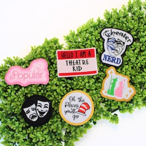 May include: A collection of embroidered patches with theater-themed designs. Patches include the words "Popular", "Hello I am a Theatre Kid", "Cheater Nerd", and "Oh the Places you'll go". The patches are in various colors and shapes.