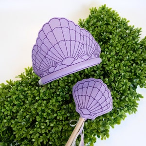 May include: Purple felt crown and wand with a seashell design. The crown is a headband with three seashells and the wand has a single seashell.