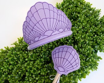 Puede incluir: Corona y varita de fieltro morado con diseño de concha. La corona es una diadema con tres conchas y la varita tiene una sola concha.