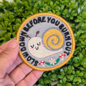 Può includere: Una toppa rotonda con sfondo grigio e bordo arancione. La toppa presenta una lumaca dei cartoni animati con guscio giallo e la scritta "SLOW DOWN BEFORE YOU BURN OUT". Piccoli funghi e fiori in basso.