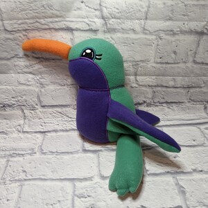 May include: A plush toy hummingbird with a green body, purple wings, and an orange beak. The toy has embroidered eyes and a whimsical expression.