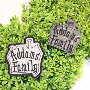Puede incluir: Dos parches bordados y un llavero con el texto "Addams Family". Los parches y el llavero tienen forma de casa y presentan el texto en una fuente gótica. Los parches tienen un borde negro. El fondo es verde.