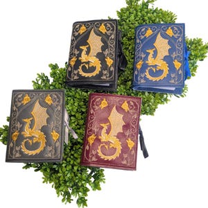 May include: Four leather-bound journals with dragon embroidery. The journals are black, blue, gray, and burgundy. Each journal features a gold dragon design with floral accents. The journals are arranged on a bed of green foliage.