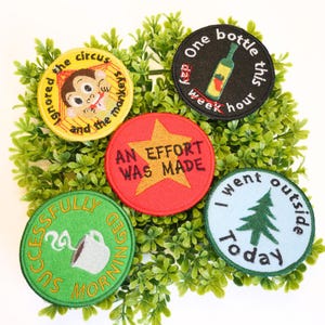 Peut inclure: Plusieurs écussons brodés avec des motifs et du texte variés. L'un représente un singe, un autre dit "An Effort Was Made", et d'autres incluent des phrases comme "One bottle this hour" et "I went outside Today."