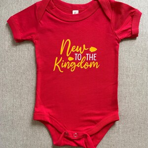 May include: A red baby bodysuit with short sleeves. The text "New to the Kingdom" is printed in yellow script across the front. The bodysuit has a snap closure at the bottom.