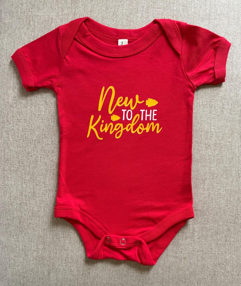 May include: A red baby bodysuit with short sleeves. The text "New to the Kingdom" is printed in yellow script across the front. The bodysuit has a snap closure at the bottom.