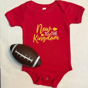 May include: A red baby onesie with the text "New to the Kingdom" in yellow. A brown and white football is next to the onesie. The onesie has snap closures.