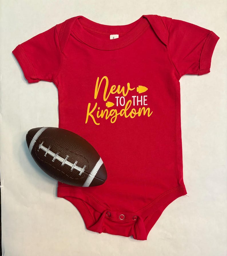 May include: A red baby onesie with the text "New to the Kingdom" in yellow. A brown and white football is next to the onesie. The onesie has snap closures.