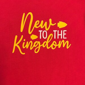 May include: A red t-shirt with the words "New to the Kingdom" in yellow script and white block letters. The design includes yellow arrowhead graphics. The shirt is a solid color.